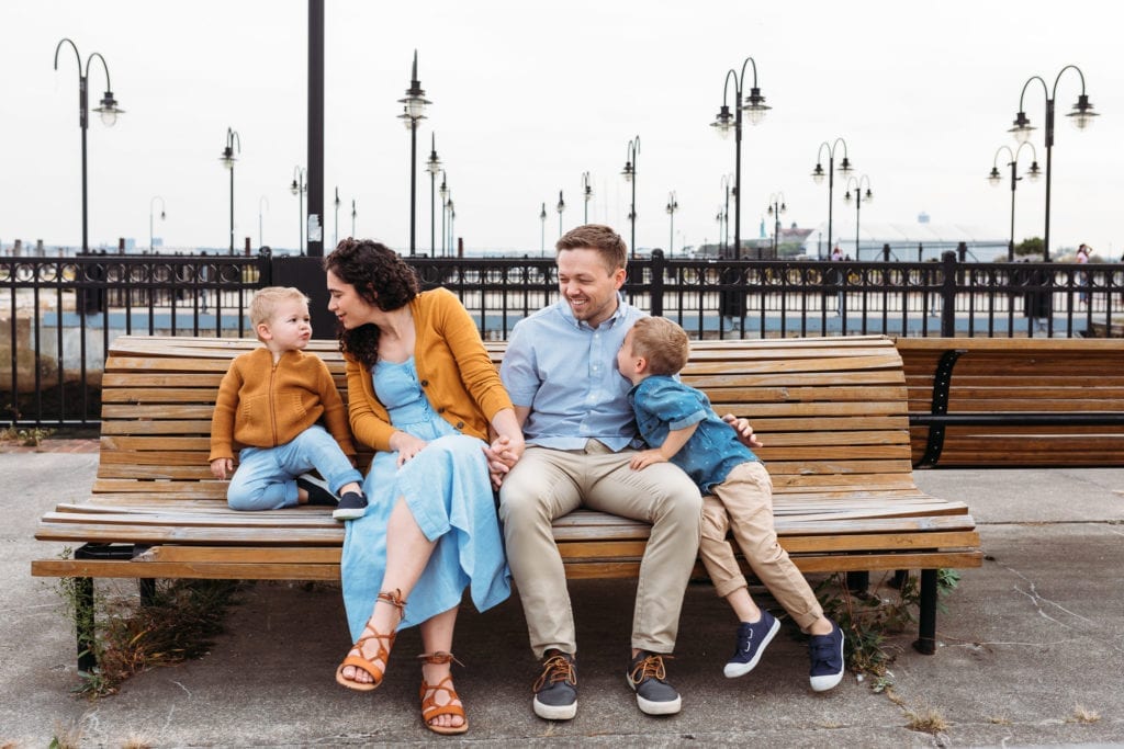 Siblings are kissing their mom and dad on a park bench at a family photo session in Liberty State Park, NJ