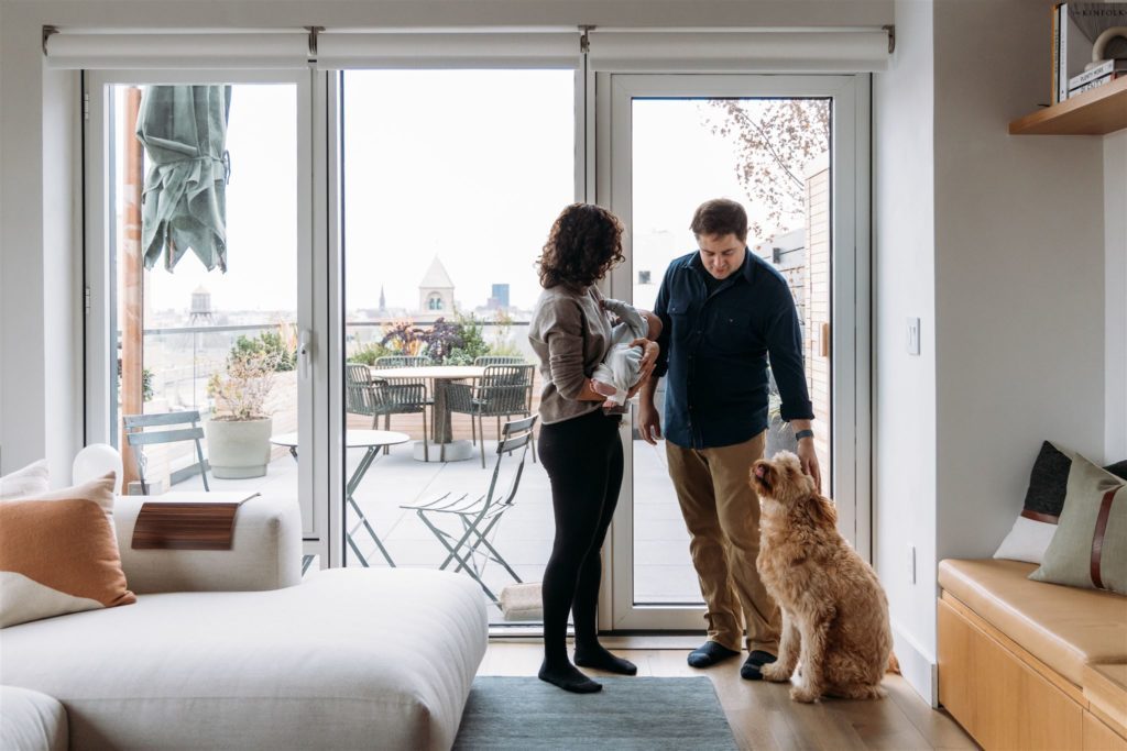 Mother, father, newborn baby and dog posing in front of a glass door leading to their NYC terrace