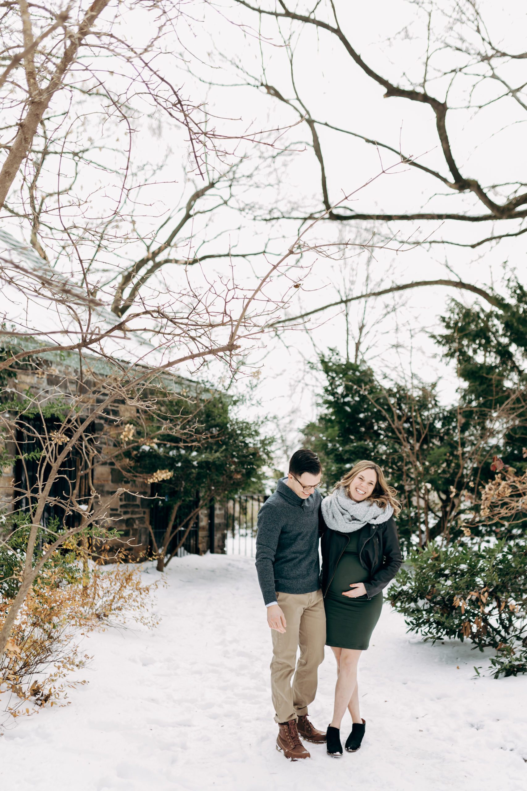 Winter couples maternity session in New York City Westchester County as a couple walks down a snowy frozen path bundled up in sweaters and scarves and cuddling close to stay warm